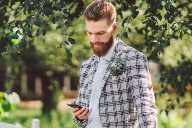 elinde telefon ile güzel saç kesimi ile seksi sakallı adam damat. Damalı ceket ve sakal şık bir saç kesimi ile Model güneşli havalarda ahşap, park ve orman bir arka plan üzerinde bir telefon kullanır