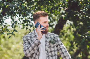 elinde telefon ile güzel saç kesimi ile seksi sakallı adam damat. Damalı ceket ve sakal şık bir saç kesimi ile Model güneşli havalarda ahşap, park ve orman bir arka plan üzerinde bir telefon kullanır