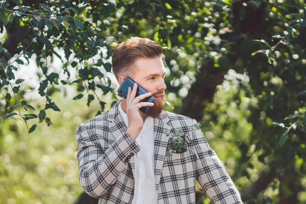 elinde telefon ile güzel saç kesimi ile seksi sakallı adam damat. Damalı ceket ve sakal şık bir saç kesimi ile Model güneşli havalarda ahşap, park ve orman bir arka plan üzerinde bir telefon kullanır