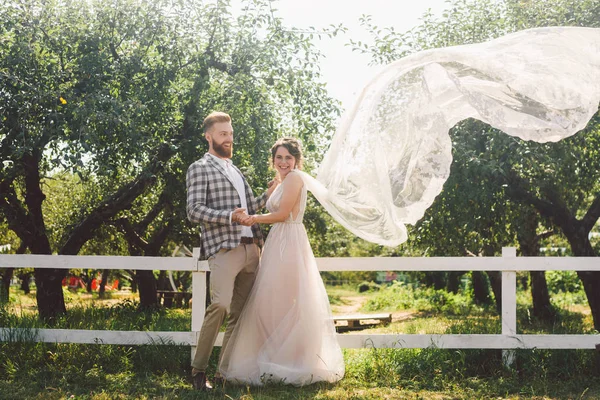 Couple caucasien amoureux mariée et marié debout dans embrasser près de bois blanc, clôture rurale dans le parc d'un verger de pommes. thème est portrait de mariage et belle robe blanche de mariage avec long voile — Photo