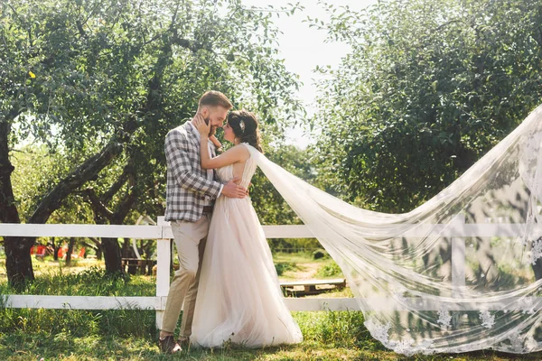 Kavkazský pár v lásce nevěsta a ženich, stojící u dřevěného bílého, venkovského plotu v parku, je jablečný sad. téma je svatební portrét a krásné svatební šaty s dlouhým závojem — Stock fotografie