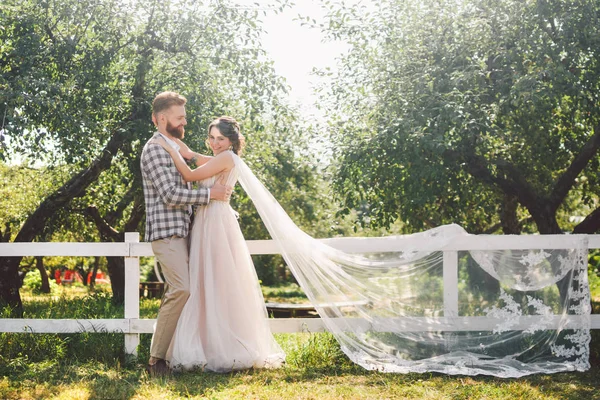 Caucasian couple in love bride and groom standing in embrace near wooden white, rural fence in park an apple orchard. theme is wedding portrait and beautiful wedding white dress with long veil