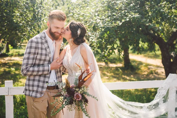 Aşık beyaz çift gelin ve damat parkta ahşap beyaz, kırsal çit yanında kucaklama ayakta bir elma bahçesi. tema uzun peçe ile düğün portre ve güzel düğün beyaz elbise