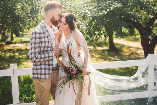 Aşık beyaz çift gelin ve damat parkta ahşap beyaz, kırsal çit yanında kucaklama ayakta bir elma bahçesi. tema uzun peçe ile düğün portre ve güzel düğün beyaz elbise