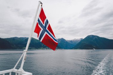 Norveç bayrağı geminin korkuluklarında sallanıyor ve suyun üzerinde sallanıyor. Bayrağı olan Norveç fiyordu. Norveç 'e feribot yolculuğu. Denizde ve dağlarda Norveç Bayrağı