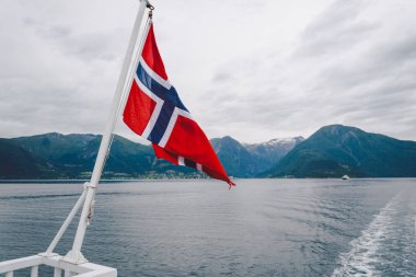 Norveç bayrağı geminin korkuluklarında sallanıyor ve suyun üzerinde sallanıyor. Bayrağı olan Norveç fiyordu. Norveç 'e feribot yolculuğu. Denizde ve dağlarda Norveç Bayrağı