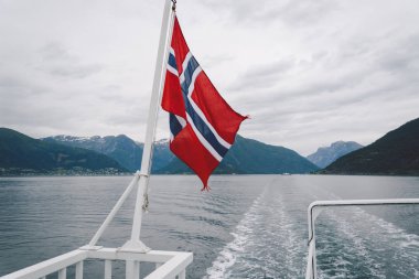 Norveç bayrağı geminin korkuluklarında sallanıyor ve suyun üzerinde sallanıyor. Bayrağı olan Norveç fiyordu. Norveç 'e feribot yolculuğu. Denizde ve dağlarda Norveç Bayrağı