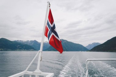 Norveç bayrağı geminin korkuluklarında sallanıyor ve suyun üzerinde sallanıyor. Bayrağı olan Norveç fiyordu. Norveç 'e feribot yolculuğu. Denizde ve dağlarda Norveç Bayrağı