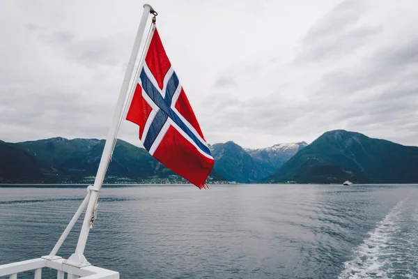 Norweska flaga wisząca na balustradzie statku i machająca nad wodą. Norweski fiord z flagą. Prom w Norwegii. Flaga Norwegii na tle morza i gór — Darmowe zdjęcie stockowe