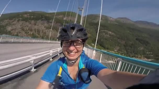 Tema de ciclismo de montaña en Escandinavia. turista humano en casco y ropa deportiva en bicicleta en Noruega en el puente colgante Hardanger Bridge lanzado a través del fiordo Hardanger en el suroeste de Noruega — Vídeo de stock