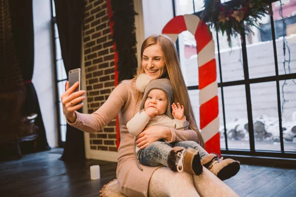 Navidad, tecnología y concepto familiar, madre feliz e hijo pequeño tomando teléfono inteligente selfie en casa. El niño y mamá hacen selfie en el teléfono móvil. madre e hijo hace foto autorretrato en invierno — Foto de Stock