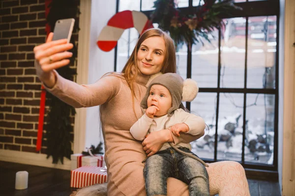 Navidad, tecnología y concepto familiar, madre feliz e hijo pequeño tomando teléfono inteligente selfie en casa. El niño y mamá hacen selfie en el teléfono móvil. madre e hijo hace foto autorretrato en invierno — Foto de Stock