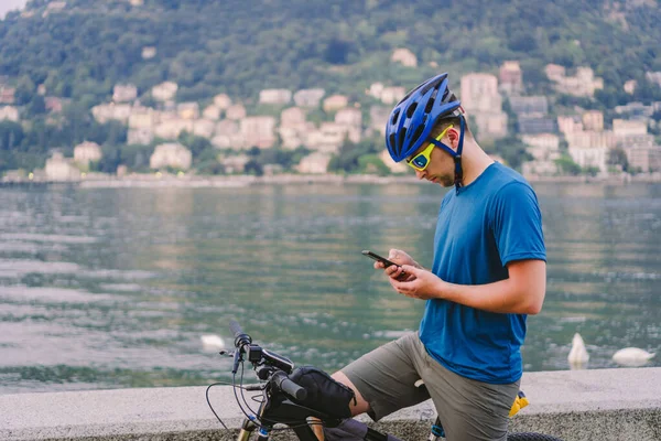 The theme of tourism and travel in Italy. A male cyclist uses a phone on the shore of Lake Como. Tourist guy in a helmet with a bicycle on the shore of a mountain lake in northern Italy