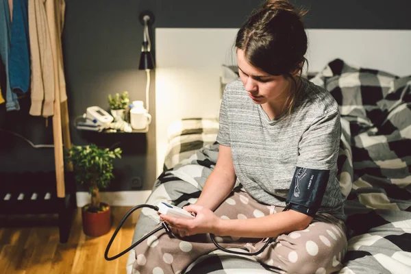 El tema de la presión arterial alta es la hipertensión. 22 años joven mujer caucásica utiliza un tonómetro automático para comprobar la presión en casa en el dormitorio en la cama en la ropa de casa por la noche — Foto de Stock