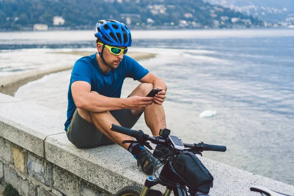 İtalya 'da turizm ve seyahat teması. Erkek bir bisikletçi Como Gölü kıyısında bir telefon kullanıyor. Kuzey İtalya 'da bir dağ gölünün kıyısında kasklı bisikletli bir turist. — Stok fotoğraf