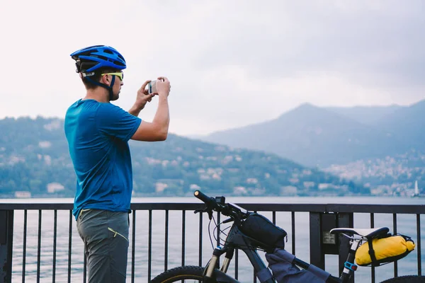 Bisikletçi akıllı telefonla fotoğraf çekiyor. Bisiklet kasklı beyaz adam manzaranın fotoğrafını çekiyor. Açık hava aktivitesi. Turist İtalya 'daki Como Gölü' nün fotoğrafını çekti — Stok fotoğraf