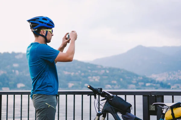 Cyclist taking pictures with smart phone. Caucasian man in bike helmet taking photo of landscape. Outdoor activity. Tourist makes photo of Lake Como in Italy