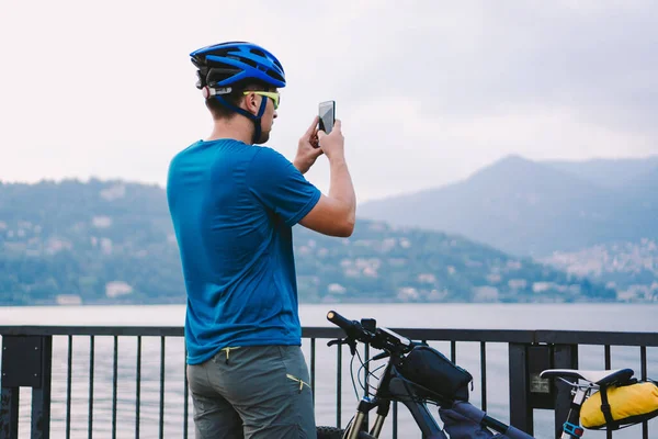 Bisikletçi akıllı telefonla fotoğraf çekiyor. Bisiklet kasklı beyaz adam manzaranın fotoğrafını çekiyor. Açık hava aktivitesi. Turist İtalya 'daki Como Gölü' nün fotoğrafını çekti — Stok fotoğraf
