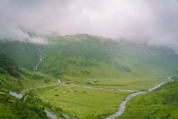 Peisaj frumos și priveliște peisagistică a Norvegiei, dealuri verzi și munte într-o zi tulbure. peisaj verde de dealuri și munte parțial acoperit cu ceață. Fermă și cabane pe un râu de ghețar — Fotografie de stoc gratuită