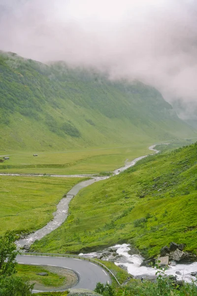 Hermoso paisaje y vista panorámica de Noruega, verdes colinas y montañas en un día nublado. paisaje verde de colinas y montañas parcialmente cubiertas de niebla. Granja y cabañas en un río glaciar — Foto de stock gratuita