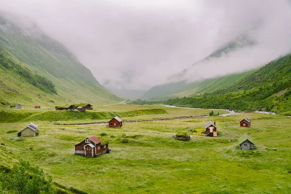 Hermoso paisaje y vista panorámica de Noruega, verdes colinas y montañas en un día nublado. paisaje verde de colinas y montañas parcialmente cubiertas de niebla. Granja y cabañas en un río glaciar — Foto de Stock