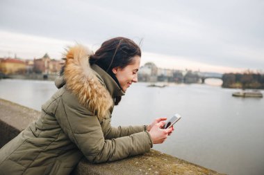 Prag 'da nehir kenarında bekleyen güzel bir kadın, kısa mesajlar. Prag Köprüsü geçmişi olan nehir kenarındaki telefona bakan bir kız. Prag şehrinde Vltava nehrinin kenarında oturan akıllı telefonlu kadın.