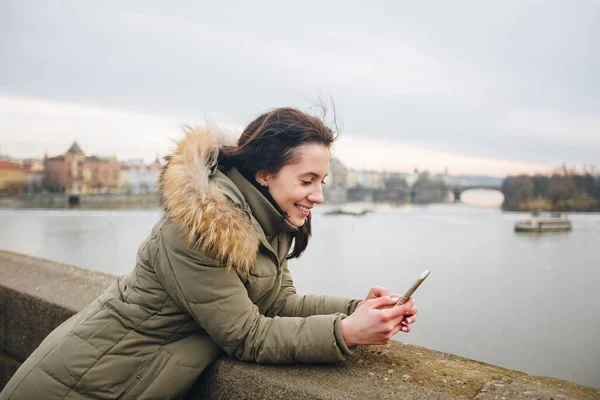 Mulher bonita em pé junto ao rio na cidade Praga, mensagens de texto. Menina olhando para o telefone na margem do rio, com fundo da ponte de Praga. Mulher com Smartphone sentado perto do rio Vltava em Praga Cidade — Fotografia de Stock
