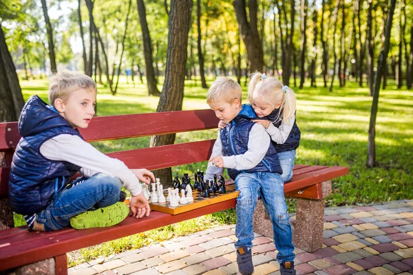 Caucasian children play chess on wooden chessboard in park bench. Brothers and sister big friendly active mental family spend time on vacation in sunny weather. Children\'s chess club. Chess sport.