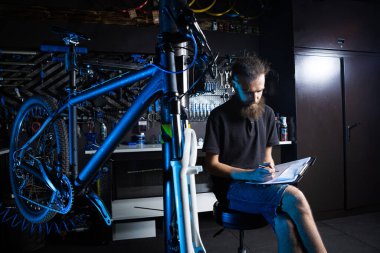 Şık bisiklet tamircisi, küçük işletme sahibi, mağaza müdürü, programını kontrol eden, not defteri ve kalem kullanan, bisiklet mağazasında yapılacak tamir işlerini not eden. Bir Bisiklet Dükkanında Kontrol Sayacı.