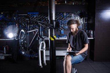 Sakallı şık bir adam, bisiklet tamircisi not alıyor, bisiklet tamirhanesinde bisiklet tamir ve onarım öncesi kontrol listesi tutuyor. Bisiklet dükkanında panoya yazı yazan beyaz bir erkek. Bisiklet arzı değerlendiriliyor.