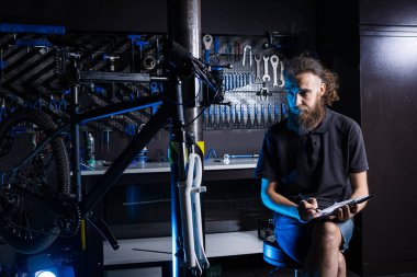 Bisiklet Atölyesi Teknisyeni İdari İşler yapıyor. Kontrol listesi defterine bisiklet tamircisi yazıyormuş. İş yerinde bisiklet tamircisi. Bisiklet dükkanında beyaz işçi panoya yazı yazıyor..