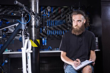 Sakallı şık bir adam, bisiklet tamircisi not alıyor, bisiklet tamirhanesinde bisiklet tamir ve onarım öncesi kontrol listesi tutuyor. Bisiklet dükkanında panoya yazı yazan beyaz bir erkek. Bisiklet arzı değerlendiriliyor.