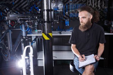 Bisiklet Atölyesi Teknisyeni İdari İşler yapıyor. Kontrol listesi defterine bisiklet tamircisi yazıyormuş. İş yerinde bisiklet tamircisi. Bisiklet dükkanında beyaz işçi panoya yazı yazıyor..