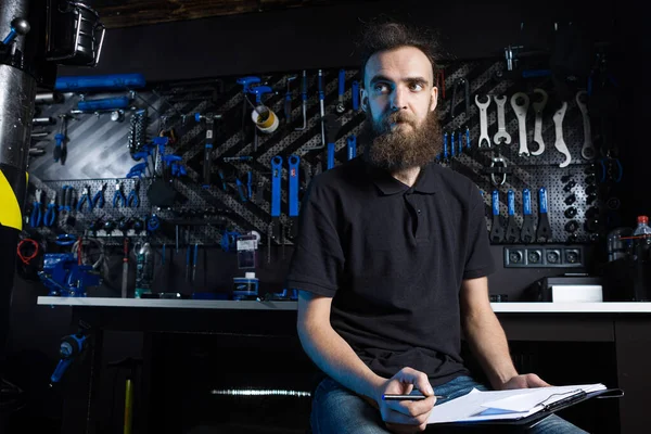 Stylish man with beard, cycle mechanic takes notes, checklist before repairing and servicing bike in bicycle shop workshop. Caucasian male in bicycle shop writing on clipboard. Assessing bike supply.