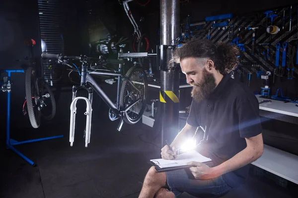 Stylish bicycle mechanic making notes in clipboard in workshop. theme small business selling, repair and service bicycles. Technician Doing Administrative paperwork in bike shop. Manage business.