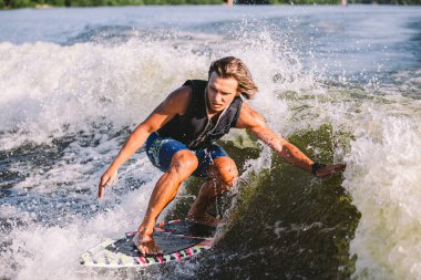 Göl dalgaları boyunca yelek giymiş güzel, uzun sarı saçlı uyanış yeleği. Atletik erkek sporcu yazın nehirde sörf yaparak uyanır. Uyanış su sporu. Dalgada sörfçü. Sörf tahtasında sörf yapan adam.