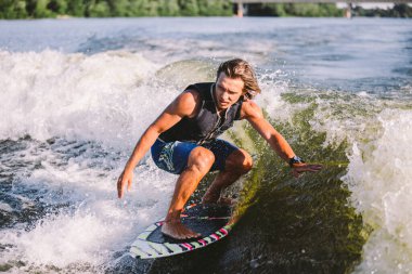 Aktif uyanış uzmanı nehir dalgalarının aşağısına atlıyor. Dalgada sörfçü. Erkek sporcu Wakesurf eğitimi alıyor. Açık havada aktif su sporları. Bir adam sörf yaparken dalga yakalar..