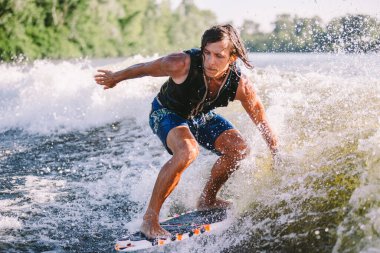 Göl dalgaları boyunca yelek giymiş güzel, uzun sarı saçlı uyanış yeleği. Atletik erkek sporcu yazın nehirde sörf yaparak uyanır. Uyanış su sporu. Dalgada sörfçü. Sörf tahtasında sörf yapan adam.