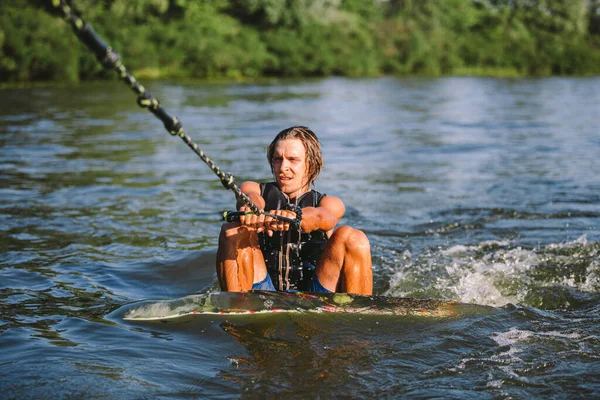 Young athletic man with long hair wakesurfing on waves of river in sunny summer weather. Ttheme outdoor activities in summer. Water sports wakesurf on the board. sliding wakeboarder in water splash.