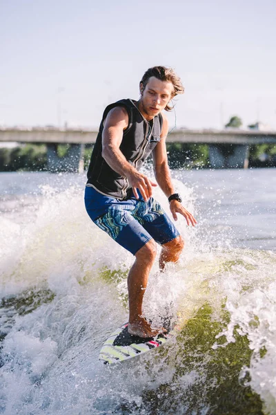 Young athletic man with long hair wakesurfing on waves of river in sunny summer weather. Ttheme outdoor activities in summer. Water sports wakesurf on the board. sliding wakeboarder in water splash.