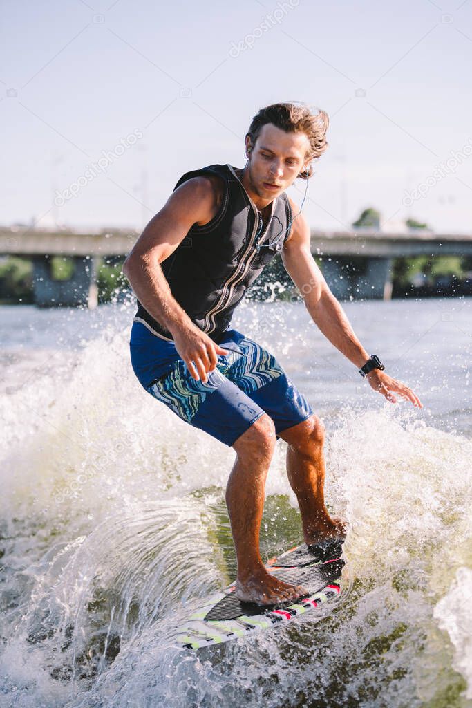 Young athletic man with long hair wakesurfing on waves of river in sunny summer weather. Ttheme outdoor activities in summer. Water sports wakesurf on the board. sliding wakeboarder in water splash.