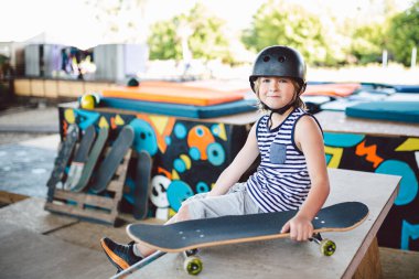 Kasklı yakışıklı beyaz bir çocuk kaykay parkındaki bir rampada oturuyor. Bir kaykaycı numaralar arasında dinleniyor. Kaykay eğitimi için atlet çocuk molası. Çocukluk ve şehir sporları teması.