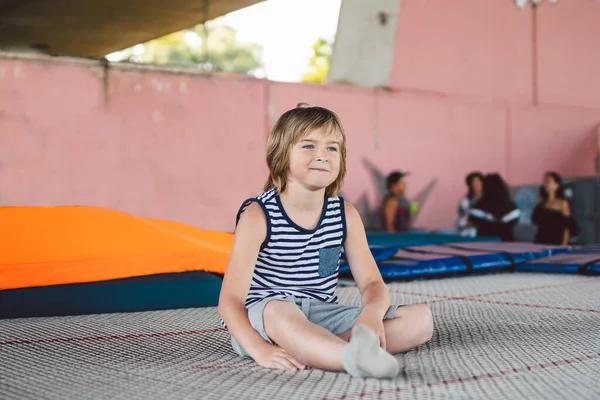 Athlete Caucasian boy sitting on trampoline after training. child is engaged in trampolining on professional trampoline outside. Little youngster relaxing from jumping on trampoline. Acrobatic sport.