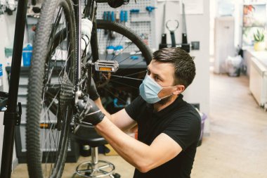 Tamirci atölyede bisiklet tamir ediyordu. Tıbbi maske takıyordu. Korunaklı yüz maskesi takan karantina altındaki koronavirüs salgınında insan bisikleti tamir edilir. İnsan döngüyü inceler ve düzeltir. Spor mağazası konsepti.