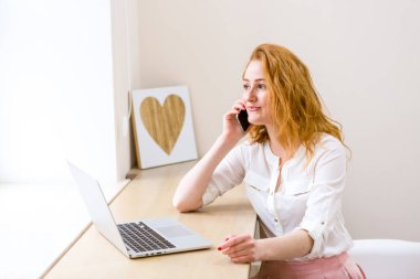 Bilgisayarlı ve akıllı telefonlu bir masada çalışan profesyonel bir iş kadını telefon görüşmesinde gülümsüyor, ofis içinde. Çalışan kadın yaşam tarzı. İş kadını Telefon görüşmesi yapıyor
