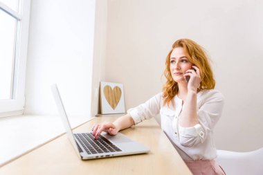 Bilgisayarlı ve akıllı telefonlu bir masada çalışan profesyonel bir iş kadını telefon görüşmesinde gülümsüyor, ofis içinde. Çalışan kadın yaşam tarzı. İş kadını Telefon görüşmesi yapıyor