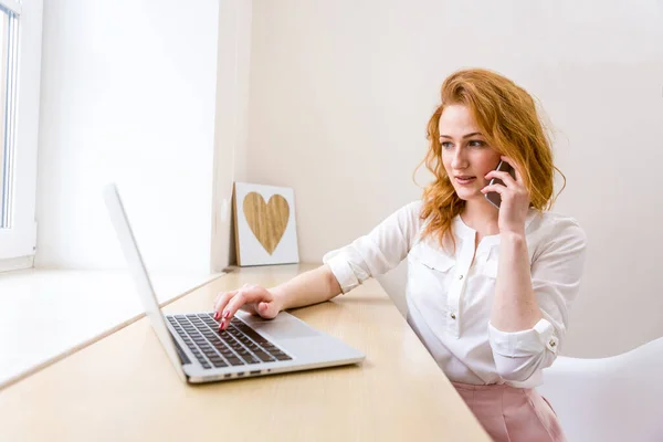 Portrait businesswoman working with laptop, type on keyboard and calling phone at office. Business woman sitting at desk in office talking with client by phone using computer giving consultation.