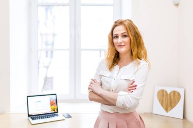 Pencerenin önünde duran genç ve güzel bir iş kadını dizüstü bilgisayarlı bir masa beyaz gömlek ve pembe etekle kollarını göğsünün üzerinde kavuşturmuş. Ofisteki iş kadını. Başarı, kariyer ve iş.