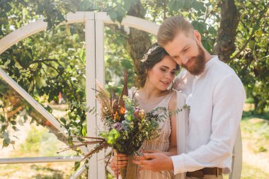 Tarz sahibi damadın düğün portresi ve bahçede parktaki şirin gelin. Güzel yeni evliler, arka planda, elma bahçesindeki ahşap düğün dekorunda şefkatle kucaklaşırlar. Çift kucaklaşması elma ağacı