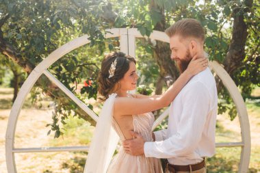 Tarz sahibi damadın düğün portresi ve bahçede parktaki şirin gelin. Güzel yeni evliler, arka planda, elma bahçesindeki ahşap düğün dekorunda şefkatle kucaklaşırlar. Çift kucaklaşması elma ağacı